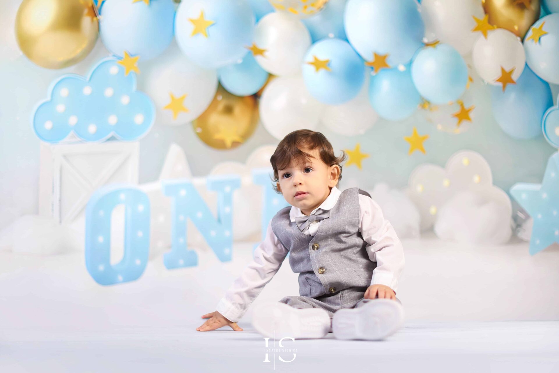 Baby boy smiling in a cloud-themed first birthday photoshoot with blue and gold balloon decor.