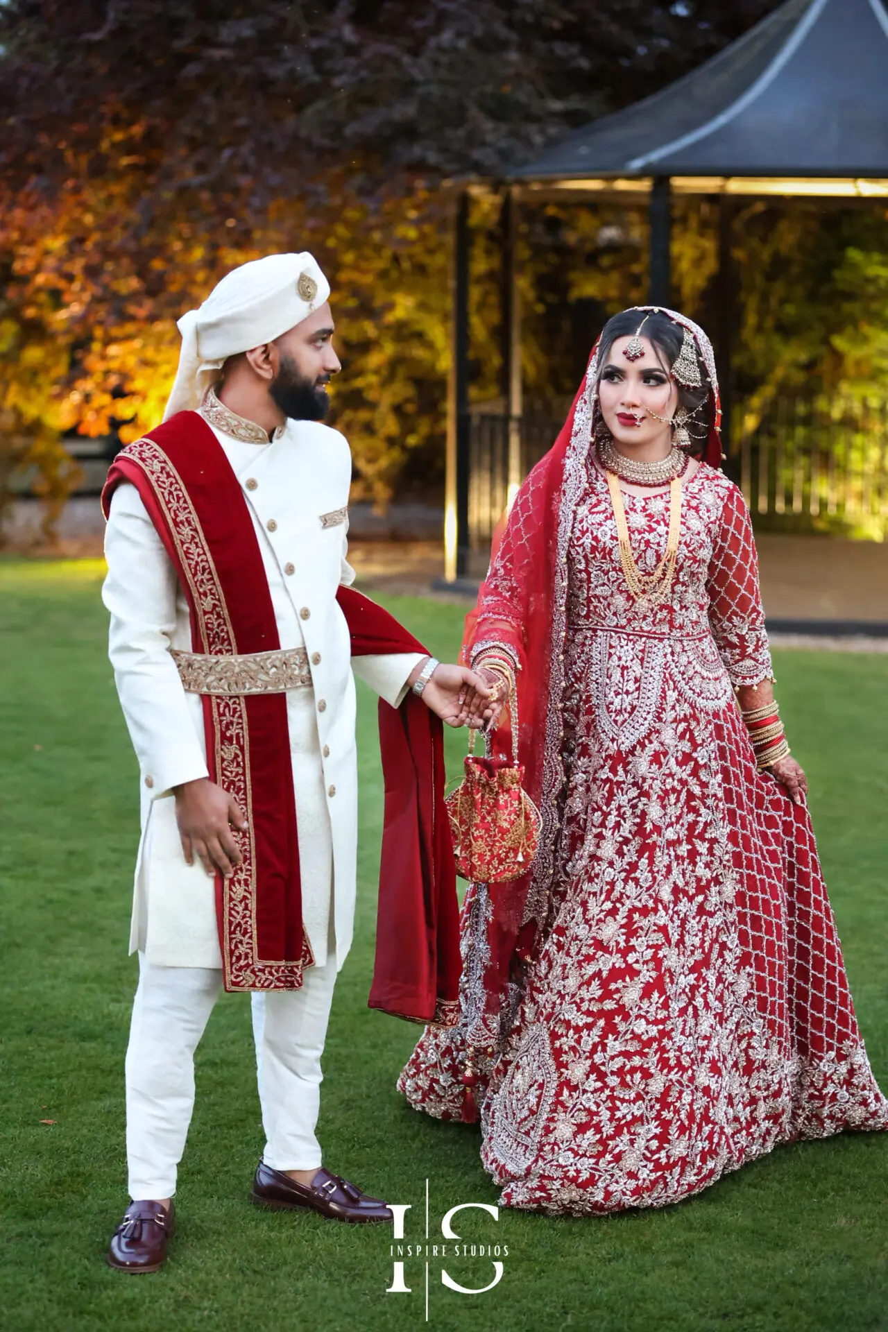 Bride and groom in traditional red and white baraat wedding attire in a garden setting in London, captured by baraat wedding photographer from Inspire Studios.