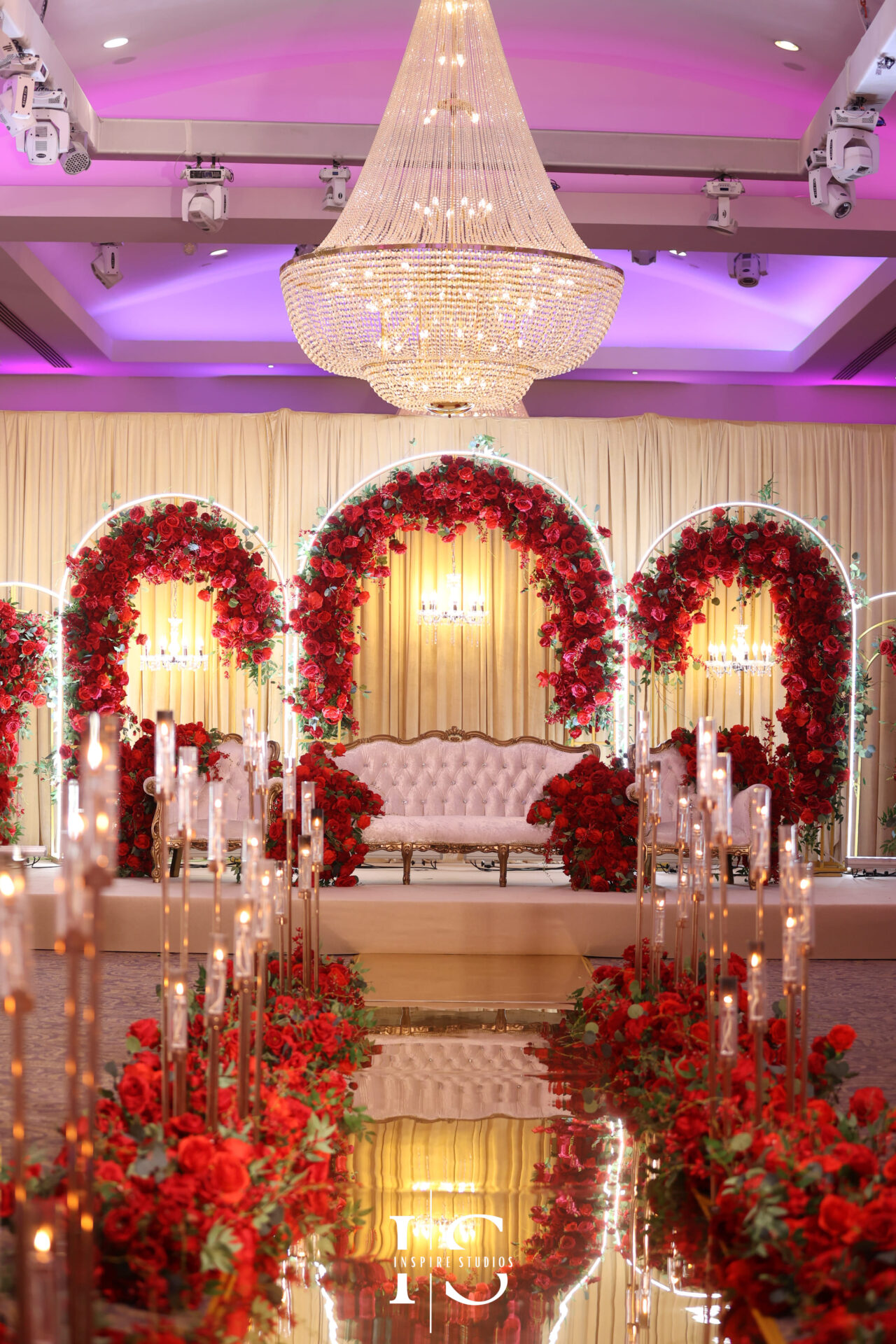 Luxurious London baraat wedding stage decorated with red roses, chandeliers, and a white tufted sofa.