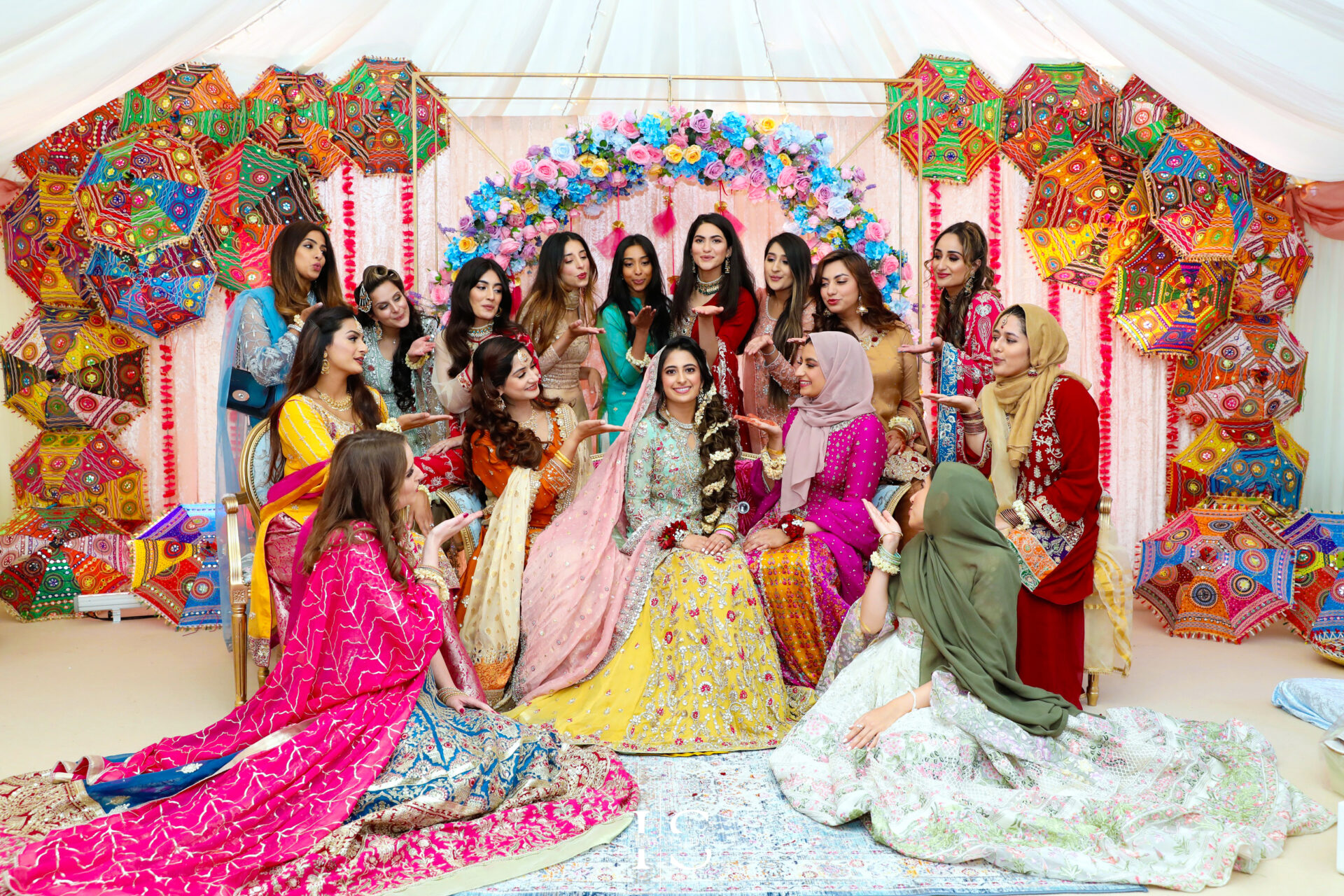 Luxury Asian wedding photography capturing a stunning bride in intricate attire and floral jewelry with bridesmaids, set against a vibrant Mehndi backdrop in London.