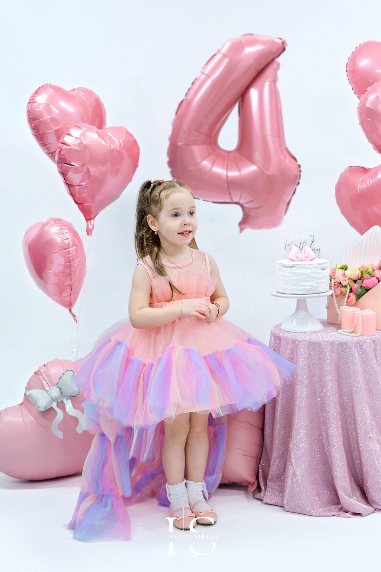 Little girl in a pastel rainbow princess dress holding pink roses during her 4th birthday photoshoot at Inspire Studios.