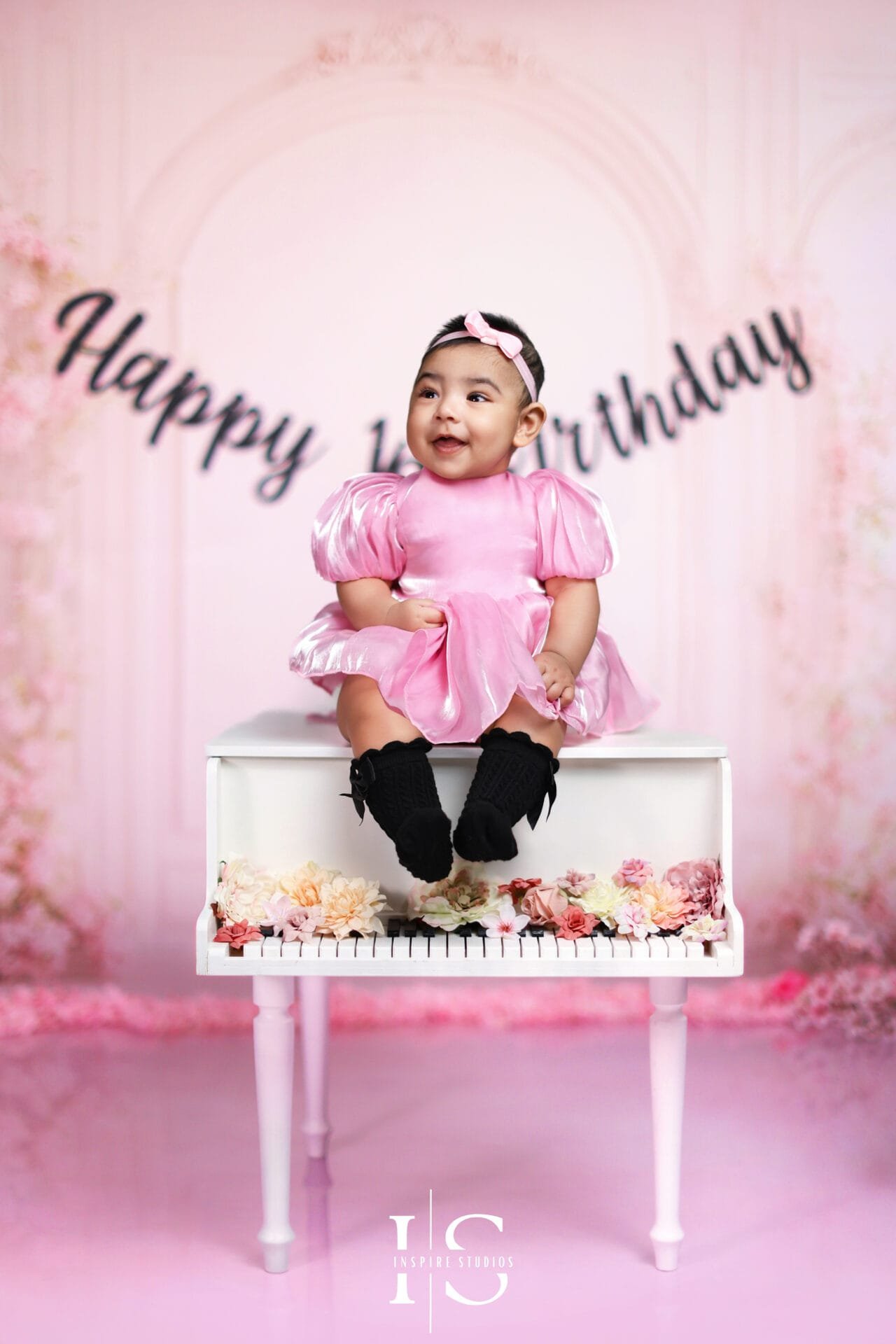 Toddler girl in a pink birthday dress sitting on a white piano during a children photoshoot at Inspire Studios.