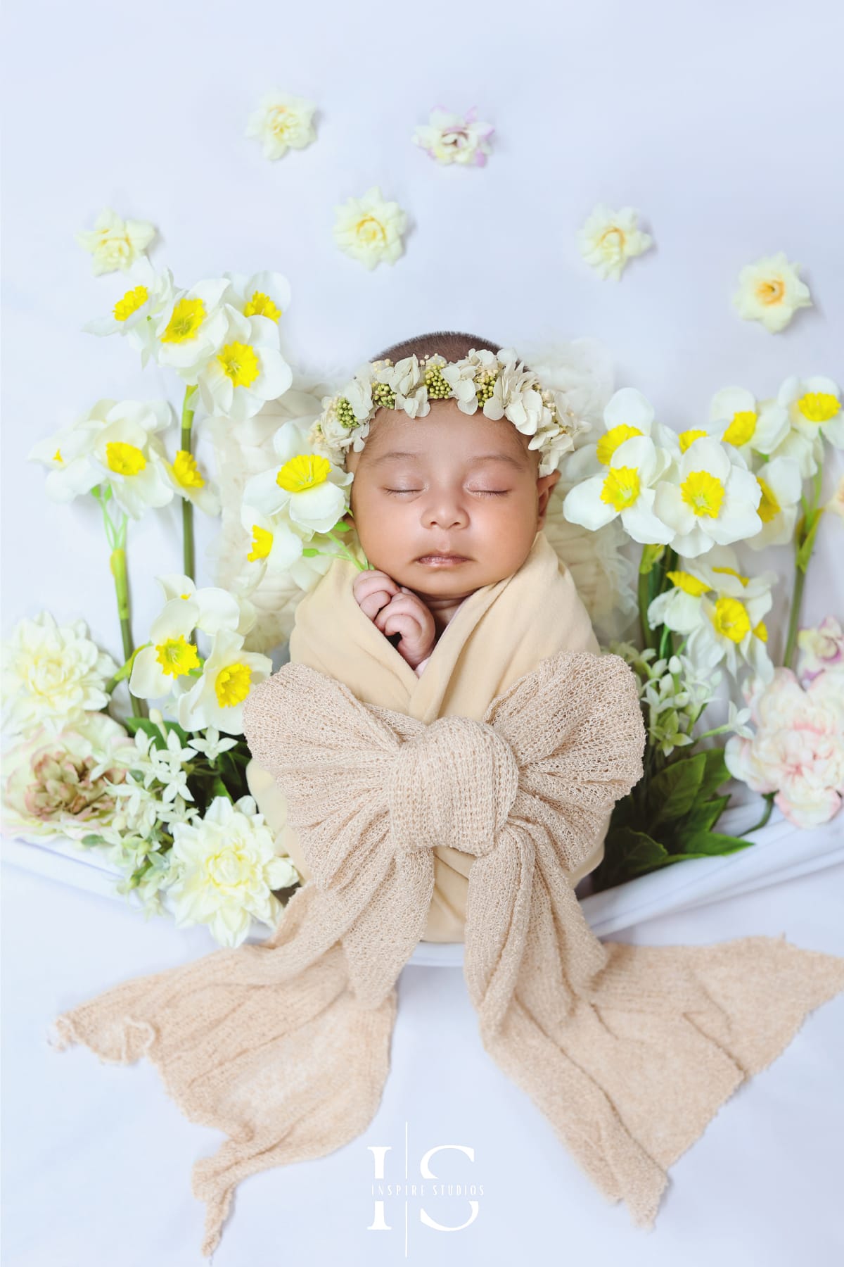 Newborn baby wrapped in a soft cream swaddle, sleeping peacefully among white and yellow daffodils in a spring-themed newborn photoshoot setup.