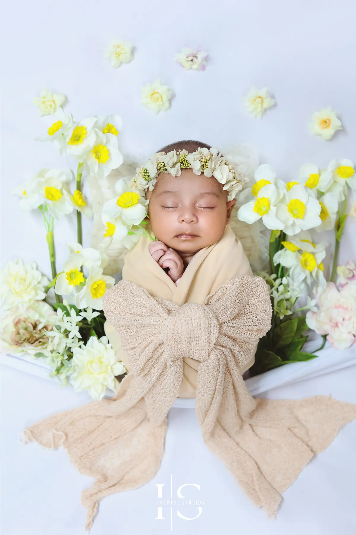 Newborn baby wrapped in a soft cream swaddle, sleeping peacefully among white and yellow daffodils in a spring-themed newborn photoshoot setup.